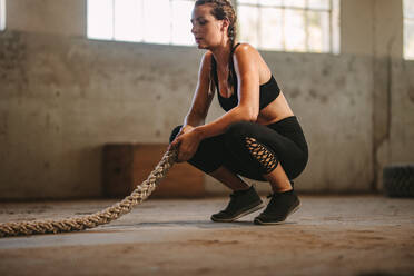 Fitness woman sitting on floor with battle rope. Woman taking rest after working out at fitness studio inside old warehouse. - JLPSF14112