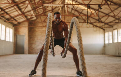 Fitness-Mann trainiert mit Kampf-Seil in Cross-Training Fitnessraum im alten Lagerhaus. Muskulöser Mann trainiert in leerem Lagerhaus. - JLPSF14116
