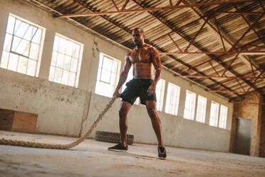 Ein fitter Mann trainiert mit Kampfseilen in einer alten Fabrik. Ein muskulöser Mann trainiert in einem Cross-Training-Fitnessstudio. - JLPSF14119