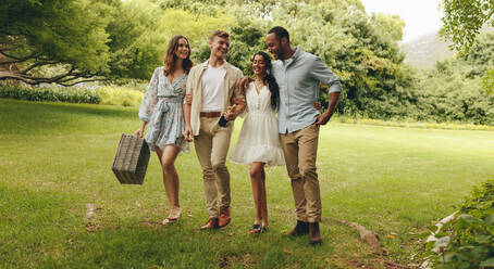 Gruppe von Freunden, die im Garten spazieren gehen. Lächelnde Männer und Frauen, die im Park spazieren gehen und einen Picknickkorb tragen. - JLPSF14143