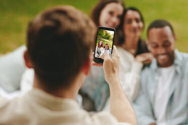 Nahaufnahme eines Mannes, der sein Handy benutzt, um Freunde beim Picknick zu fotografieren; Fokus auf das Handy-Display. - JLPSF14187