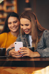 Frauen sitzen in einem Café zusammen, schauen auf ihr Handy und lächeln. Junge Frau zeigt ihrer Freundin etwas auf ihrem Smartphone. - JLPSF14190