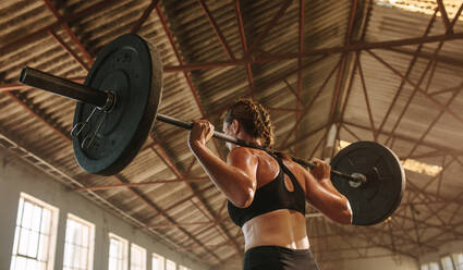 Strong woman exercising with barbell. Fit woman working out with heavy weights at cross training gym in factory shade. - JLPSF14206