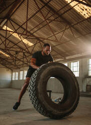Männlicher Mann beim Reifen-Weitwurf-Workout. Ein Mann wirft einen Reifen während einer intensiven Trainingseinheit in einem leeren Fabrikschatten. - JLPSF14208