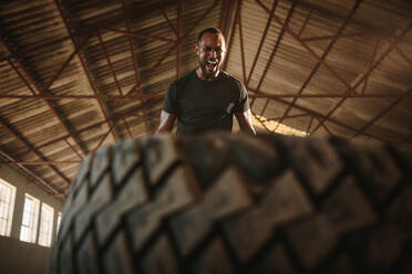 Ein Mann trainiert in einer leeren Lagerhalle mit einem Reifen. Ein starker männlicher Athlet wirft einen Reifen während einer intensiven Trainingseinheit. - JLPSF14211