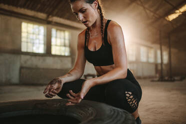 Muskulöse Sportlerin, die eine Pause vom Crosstraining einlegt. Frau in schwarzer Sportkleidung sitzt neben einem großen Reifen in einer leeren Lagerhalle. - JLPSF14215