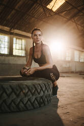 Frau sitzt nach dem Training neben einem großen Reifen. Muskulöse Frau macht eine Pause vom Crosstraining im Schatten einer alten Fabrik. - JLPSF14216