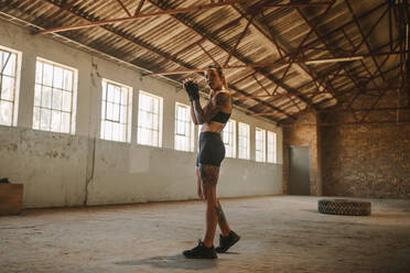 Full length of a woman in sportswear standing inside old factory shade. Fitness woman exercising shadow boxing in empty warehouse. - JLPSF14220