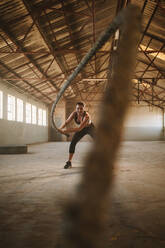 Fitness woman exercising with battle rope in cross training gym inside old warehouse. Female athlete working out at empty warehouse. - JLPSF14223