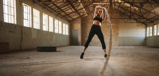 Fitness woman workout out with battling rope at cross training gym. Tough woman working out inside old warehouse. - JLPSF14224