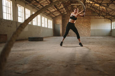 Strong woman moving battle rope in wave pattern in cross training gym. Woman doing battling rope workout in old warehouse. - JLPSF14225