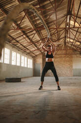 Sportlerin beim Training mit einem Battle Rope in einem alten Lagerhaus. Starke Frau beim Training mit Battle Rope im Fitnessstudio. - JLPSF14226