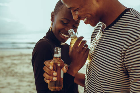 Schönes afrikanisches Paar verbringt schöne Zeit zusammen am Strand. Lächelnder Mann und Frau stehen am Strand mit Bier. - JLPSF14263