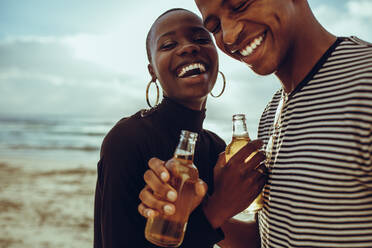 Afrikanisches Paar im Urlaub bei einem Bier am Strand. Fröhlicher junger Mann und Frau im Sommerurlaub bei einem Bier und lächelnd am Strand. - JLPSF14264
