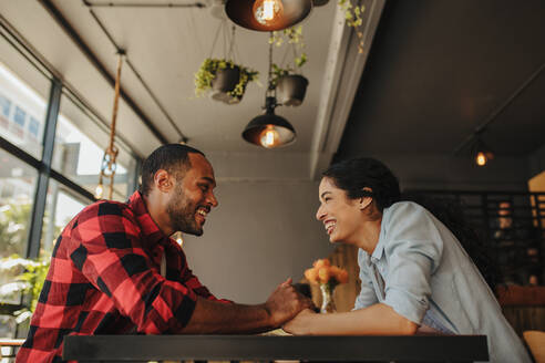 Ein fröhliches Paar sitzt im Café und hält sich an den Händen. Ein Mann und eine Frau sitzen an einem Restauranttisch und schauen sich lächelnd an. - JLPSF14303