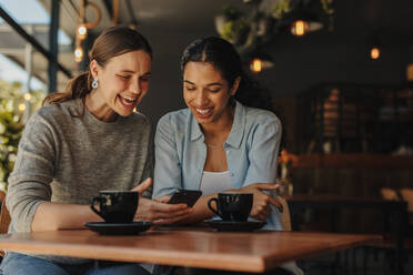 Zwei Frauen sitzen in einem Café und lächeln, während sie ein Smartphone benutzen. - JLPSF14314