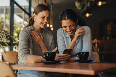 Zwei Freundinnen sitzen an einem Tisch in einem Café und schauen auf ein Handy und lächeln. Eine Frau zeigt ihrer Freundin einen lustigen Social-Media-Post auf ihrem Handy und lächelt. - JLPSF14315