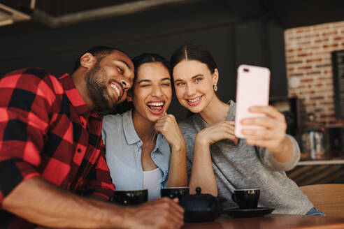 Junge Männer und Frauen, die an einem Cafétisch sitzen und ein Selbstporträt mit einem Mobiltelefon aufnehmen. - JLPSF14321