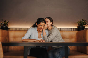 Eine Frau flüstert einer anderen Freundin ein Geheimnis ins Ohr. Zwei Freundinnen sitzen in einem Café und tratschen miteinander. - JLPSF14325