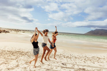 Drei Freundinnen, die sich gemeinsam am Strand vergnügen. Freunde tanzen gemeinsam am Strand und halten sich an den Händen. - JLPSF14333
