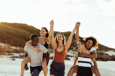 Multiethnic friends in playful mood enjoying on beach. Cheerful men and women having fun enjoying piggy riding. - JLPSF14341