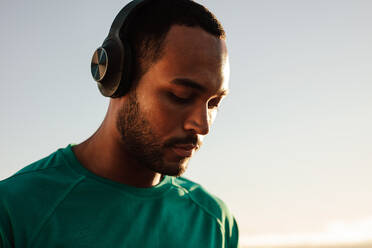 Porträt eines afroamerikanischen Sportlers, der einen kabellosen Kopfhörer trägt. Nahaufnahme eines Fitness-Mannes, der im Freien steht und Musik hört. - JLPSF14354