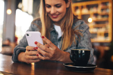 Frau sitzt in einem Café mit einer Tasse Kaffee auf dem Tisch und liest eine Textnachricht auf ihrem Smartphone. - JLPSF14357