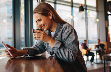 Schöne Frau schaut auf ihr Handy und trinkt Kaffee im Café. Frau in Freizeitkleidung sitzt in einem Café und schaut auf ihr Smartphone und lächelt. - JLPSF14361