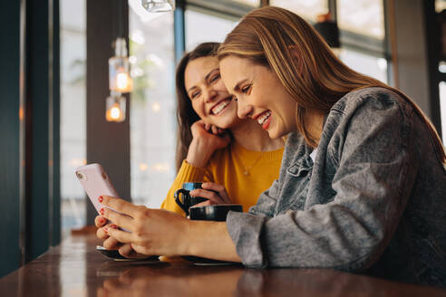 Frau mit ihrer Freundin, die an einem Kaffeetisch sitzt und auf ein Handy schaut. Glückliche Freundinnen in einem Café, die ein Handy benutzen. - JLPSF14374