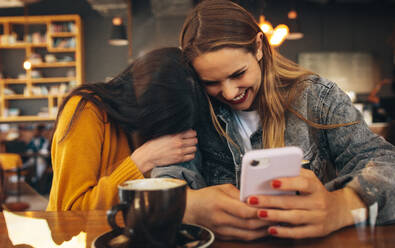 Fröhliche junge Frauen mit Smartphone und einer Tasse Kaffee auf dem Tisch. Freundinnen im Café lachen, nachdem sie sich einige lustige Bilder auf dem Handy angesehen haben. - JLPSF14376