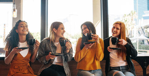 Female friends sitting in cafe holding a cup of coffee and smiling. Group of four women having coffee together at a coffee shop. - JLPSF14377