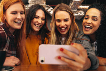 Multirassische Gruppe von Freunden, die ein Selfie mit ihrem Smartphone in einem Café machen. Junge Frauen, die am Cafétisch sitzen und ein Selbstporträt mit ihrem Handy machen. - JLPSF14382