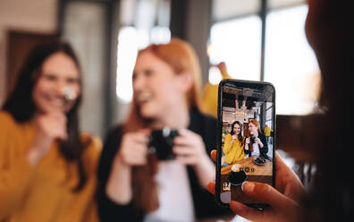 Woman sitting at a restaurant taking photograph of her friends having coffee. Woman shooting pictures with mobile phone of female friends at cafe. - JLPSF14390