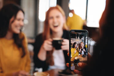 Woman taking photo of her friends with her smart phone at a coffee shop. Focus on mobile phone screen. Female friends having coffee being photographed by a women in coffee shop. - JLPSF14391