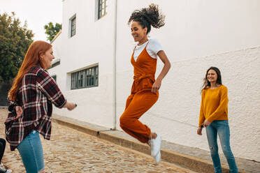 Zwei Frauen halten ein Springseil in der Hand und eine Freundin springt. Gruppe von Freundinnen, die gemeinsam Seil springen. - JLPSF14412