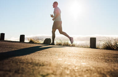 Sportler, der auf einer steilen Straße mit Sonneneinstrahlung im Hintergrund läuft. Niedriger Winkel eines Fitness-Mannes beim Laufen. - JLPSF14414