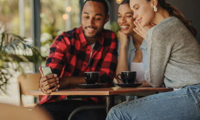 Freunde, die auf ihr Smartphone schauen, während sie in einem Café sitzen. Gemischtrassige Menschen, die an einem Tisch in einem Restaurant sitzen und ein Mobiltelefon benutzen. Fokus auf das Mobiltelefon in der Hand eines Mannes. - JLPSF14432