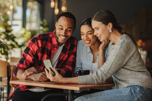 Eine Gruppe von Freunden sitzt im Café und schaut auf ihr Smartphone. Eine Frau zeigt ihren Freunden etwas auf ihrem Handy. - JLPSF14433