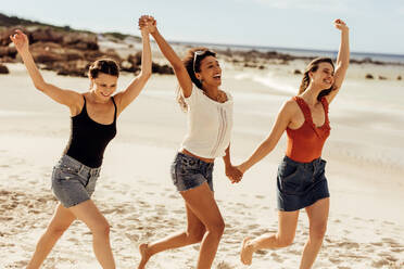 Glückliche Freundinnen, die zusammen am Strand spazieren gehen und sich an den Händen halten. Drei junge Frauen genießen und tanzen zusammen in einem Strandurlaub. - JLPSF14450