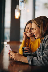 Junge Freunde, die in einem Café sitzen, auf ein Smartphone schauen und lächeln. Freunde, die in einem Restaurant sitzen, wobei eine Frau ihrem Freund etwas Interessantes zeigt. - JLPSF14467