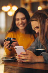 Zwei junge Frauen benutzen ein Smartphone in einem Café. Zwei lächelnde Freundinnen benutzen ein Mobiltelefon in einem Cafe - JLPSF14469