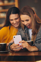 Freundinnen treffen sich in einem Café und schauen auf ihr Handy. Eine Frau zeigt einer Freundin etwas Lustiges auf ihrem Handy. - JLPSF14470