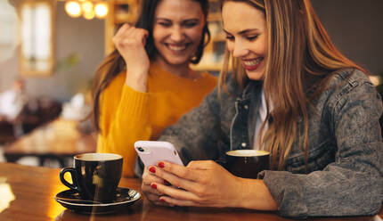 Zwei Frauen, die in einem Café sitzen, auf ein Handy schauen und lachen. Beste Freundinnen hängen in einem Café ab. - JLPSF14471