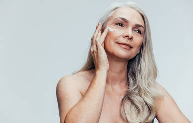 Ältere Frau, die Anti-Aging-Creme auf ihr Gesicht aufträgt und wegschaut. Frau, die Feuchtigkeitslotion auf ihr Gesicht aufträgt, vor grauem Hintergrund. - JLPSF14485