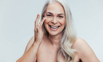 Fröhliche ältere Frau, die sich mit Feuchtigkeitscreme eincremt und lacht. Frau, die sich mit Kosmetikcreme eincremt, vor grauem Hintergrund. - JLPSF14486