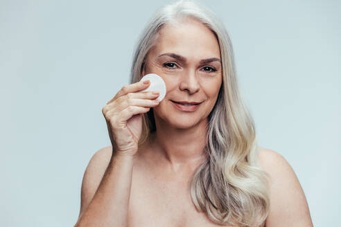 Schöne ältere Frau, die ihre Haut mit einem Wattepad reinigt. Kaukasische Frau, die ihr Gesicht mit einem Wattepad auf grauem Hintergrund reinigt. - JLPSF14488