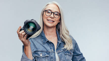 Älterer professioneller Fotograf, der eine DSLR-Kamera hält. Weiblicher Fotograf mit einer professionellen Kamera auf grauem Hintergrund. - JLPSF14509