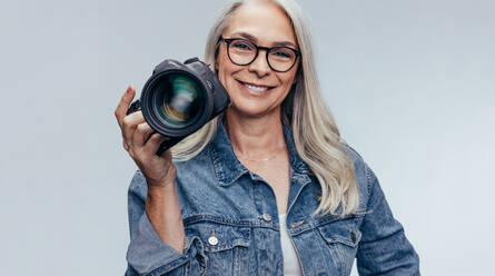 Selbstbewusste ältere Frau in Freizeitkleidung, die eine digitale Spiegelreflexkamera auf grauem Hintergrund hält. Professionelle Fotografin mit Digitalkamera. - JLPSF14510