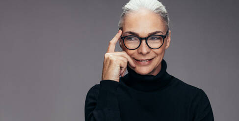 Close up of a senior woman with eyeglasses looking away and thinking against grey background. Mature woman thinking of an idea. - JLPSF14526