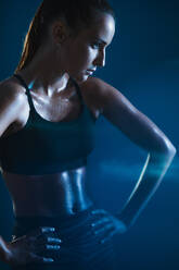 Fitte junge Frau in Sportkleidung vor blauem Hintergrund. Sportlerin mit verschwitztem Körper schaut weg. - JLPSF14571